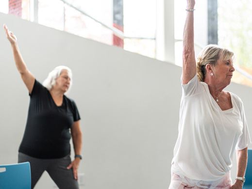 Two older adults participating in a fitness class, stretching their arms upward.