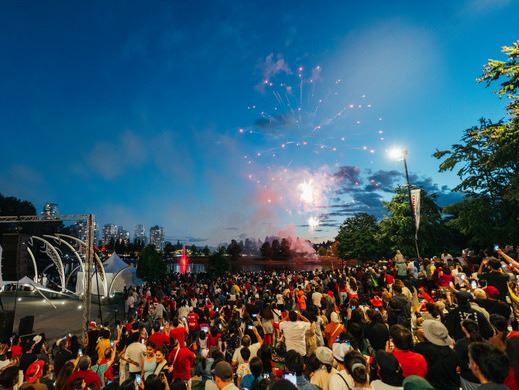 A large crowd gathers at an outdoor event during twilight to watch fireworks, with city buildings in Opens in new window