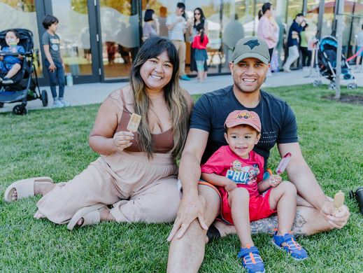 Family sitting on grass enjoying popsicles at an outdoor community event.