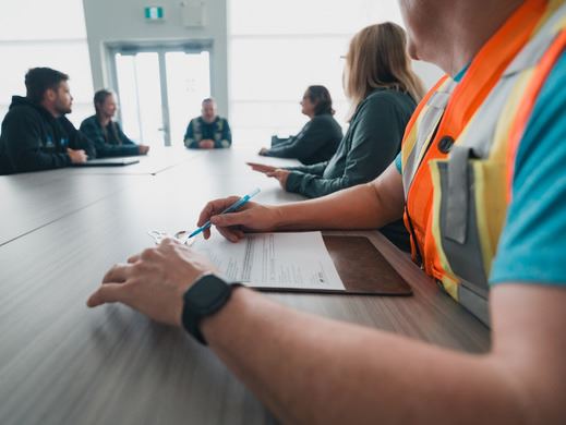 A few people sitting around a table, the foreground features a man wearing a bright safety vest.