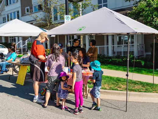A group of parents and kids socializing underneath a white tent on a neighbourhood street.