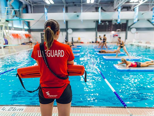 A female lifeguard wearing a red shirt that says Staff looks out over the pool full of people.